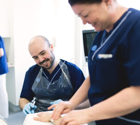 Två medarbetare tar hand om en patient som ligger i sin säng. Två medarbetare tar hand om en patient som ligger i sin säng.